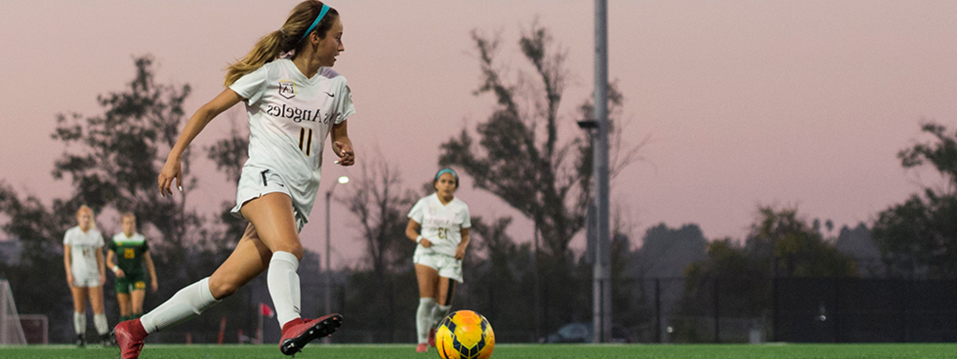 Image of a member of the  women's soccer team on the pitch at dusk, pink sky behind her. 