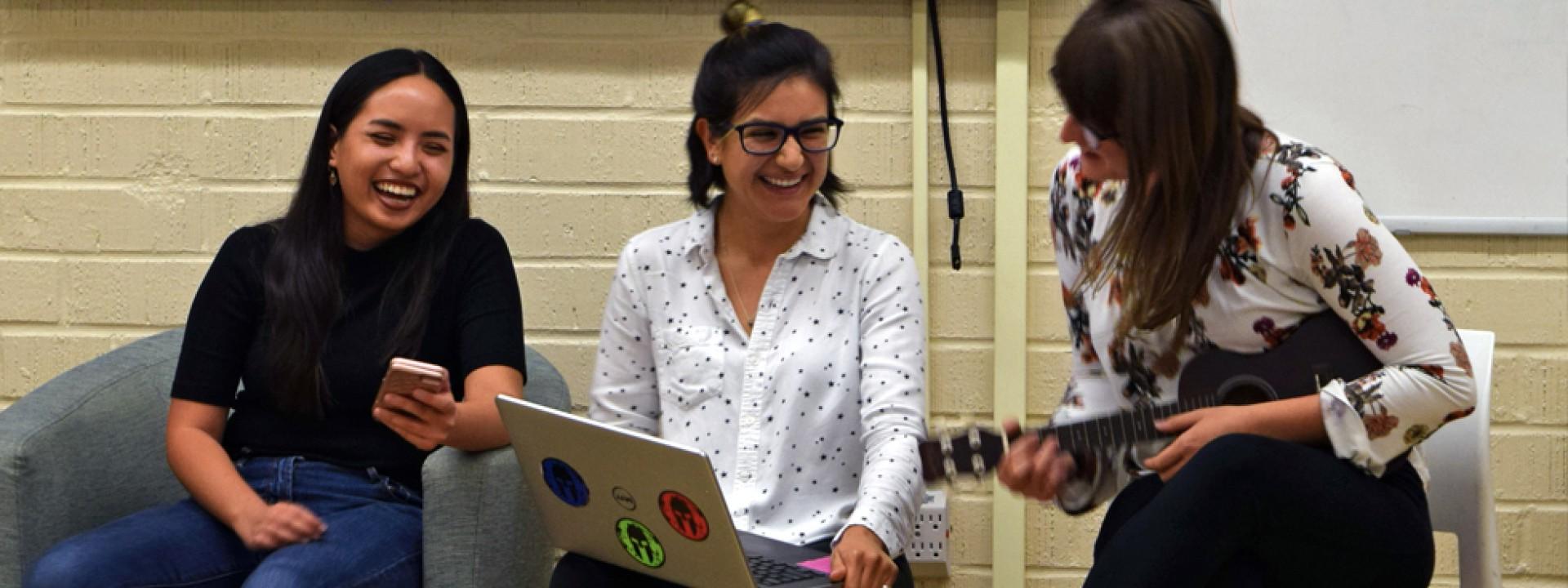 Three female students smiling, one playing a ukalele. 